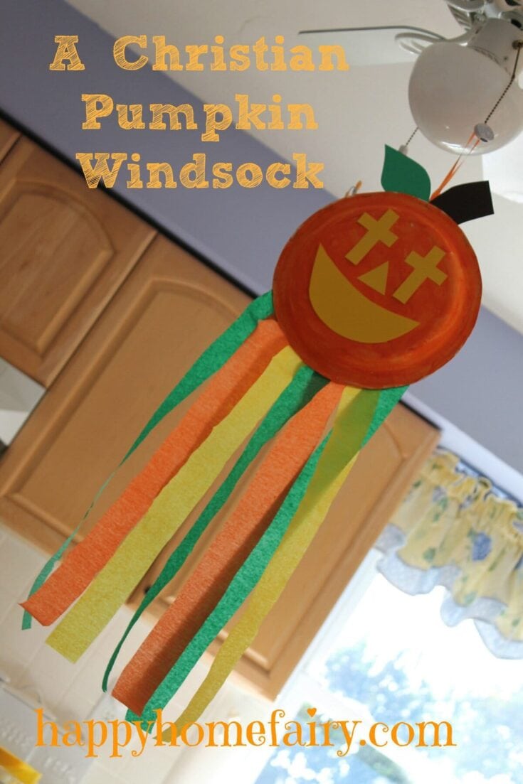 Colorful Christian-themed pumpkin windsock hanging from ceiling fan, featuring a smiling pumpkin face with a cross and rainbow-colored streamers, perfect for fall decorations.
