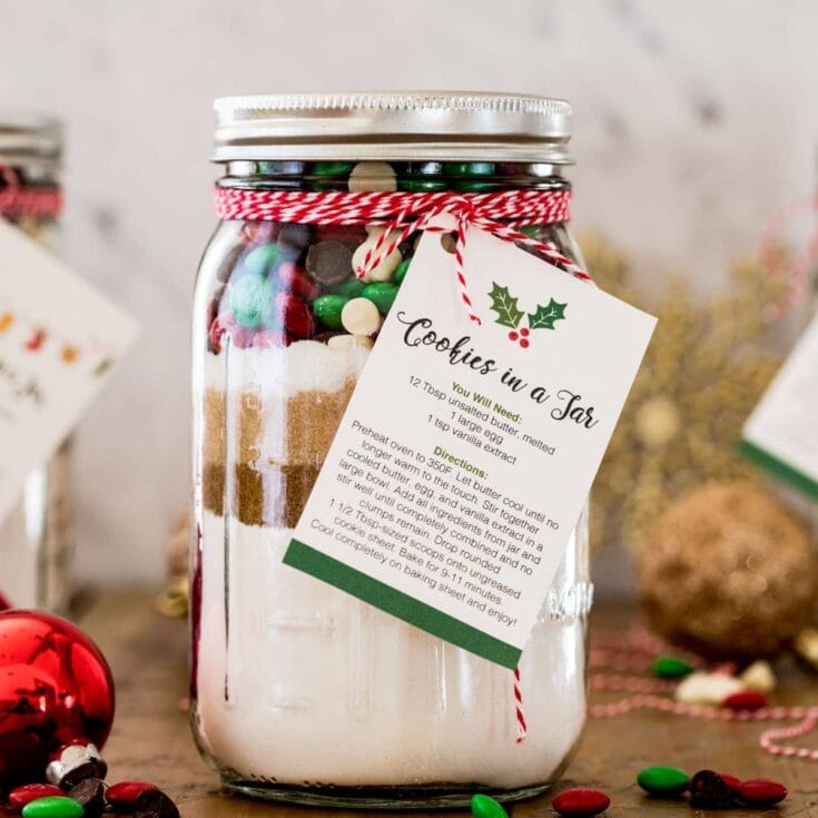 Decorative glass jar filled with layered cookie ingredients, labeled 'Cookies in a Jar' with holiday-themed tag, surrounded by Christmas ornaments and festive decorations.