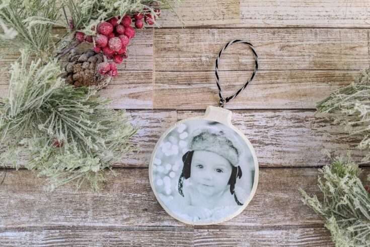 Decorative Christmas ornament featuring a black-and-white baby photo, surrounded by holiday greenery and red berries on a rustic wooden background.