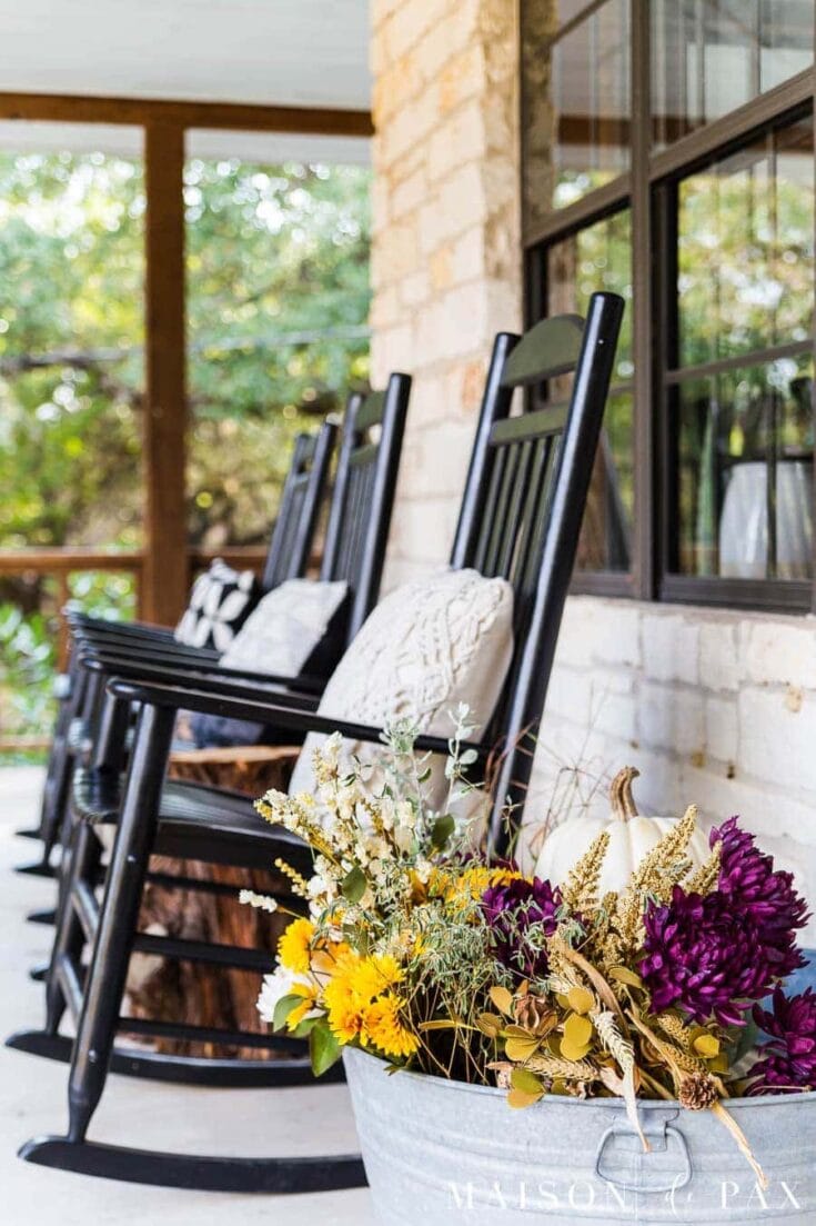 Vibrant fall flower arrangement with yellow, purple, and white blooms in a white pot, placed on a porch with black rocking chairs and a brick wall, surrounded by greenery.