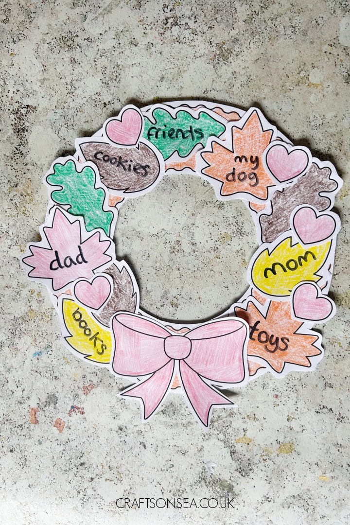 Colorful paper wreath with words like 'dad', 'mom', 'dog', 'friend', and 'cookies', decorated with pink hearts and a pink bow, on a textured surface.