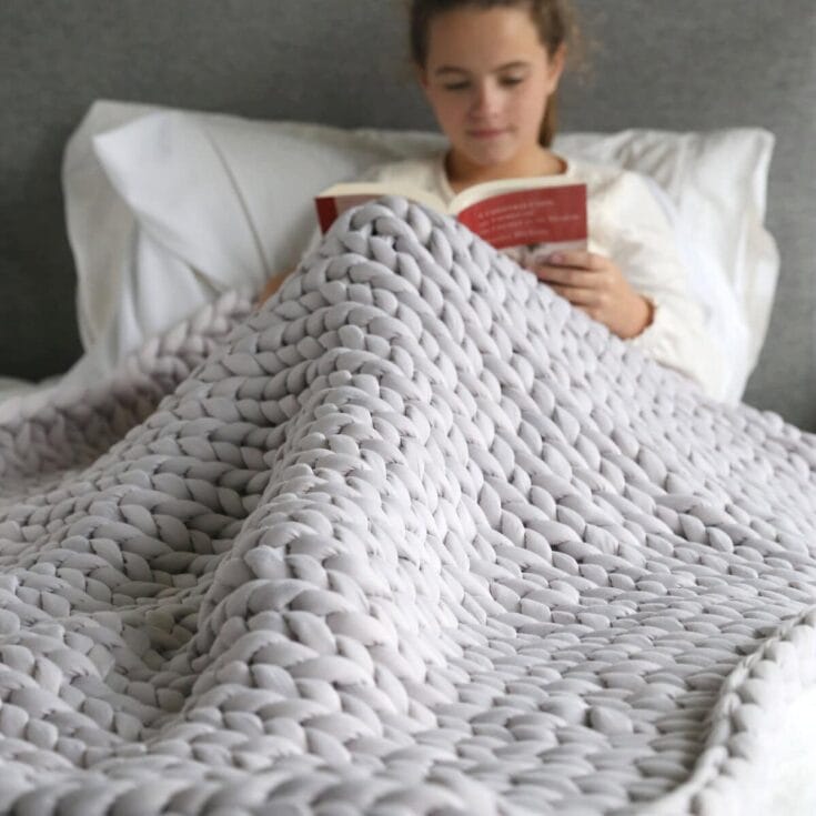 Young girl reading a book in bed covered with a large, soft, knitted gray blanket, creating a cozy and inviting atmosphere for relaxation and reading.