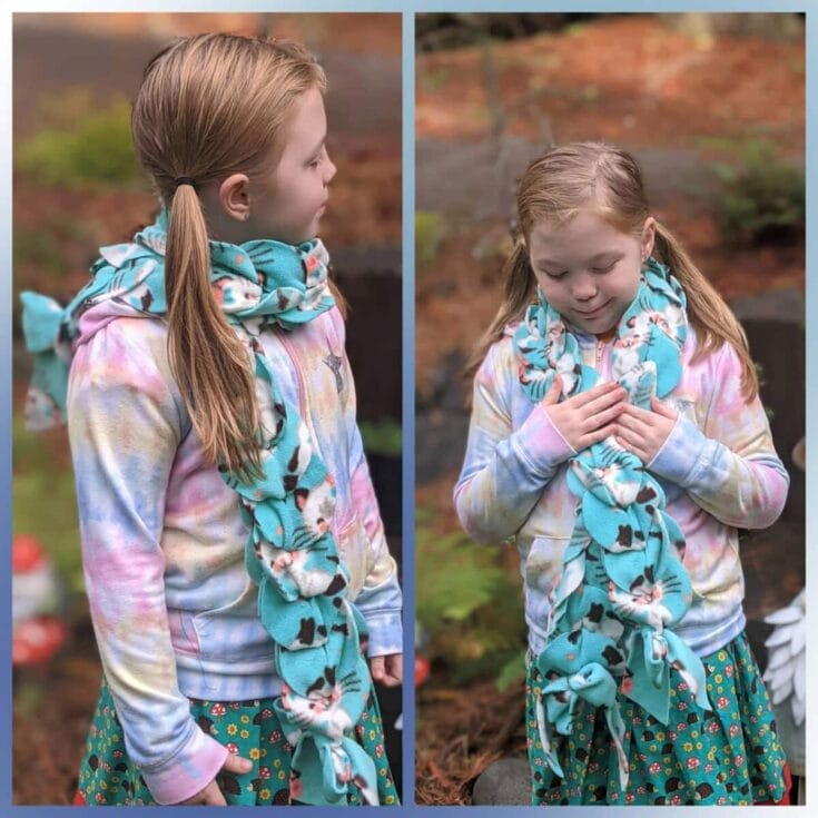 Two young girls wearing pastel-colored hoodies and vibrant skirts, each with a blue patterned scarf, standing outdoors in a garden setting with blurred greenery in the background.