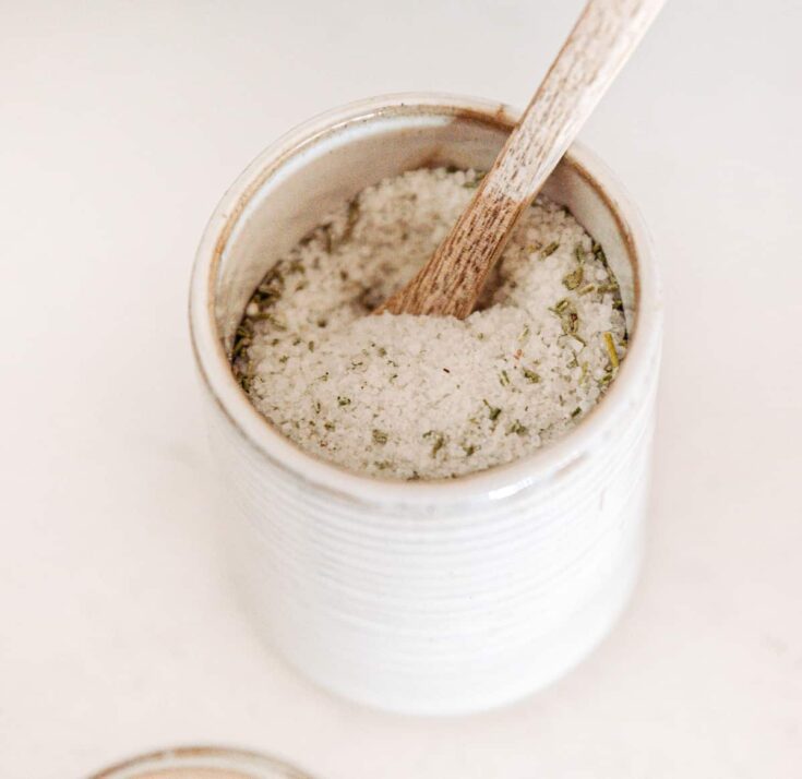 A ceramic jar filled with herbal bath salt, with a wooden spoon inside, on a white surface, ideal for relaxation and self-care.
