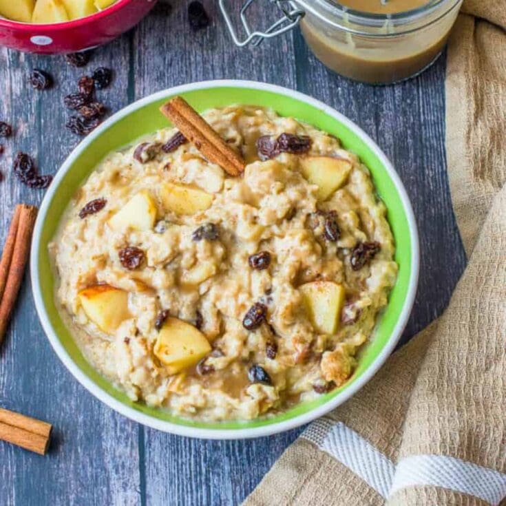 A bowl of warm apple cinnamon oatmeal topped with raisins and a cinnamon stick, served on a rustic wooden table with a beige napkin and a glass of milk.