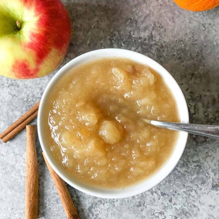 Close-up of homemade applesauce in a white bowl with apple and cinnamon sticks on a gray surface, perfect for healthy snacks and desserts.