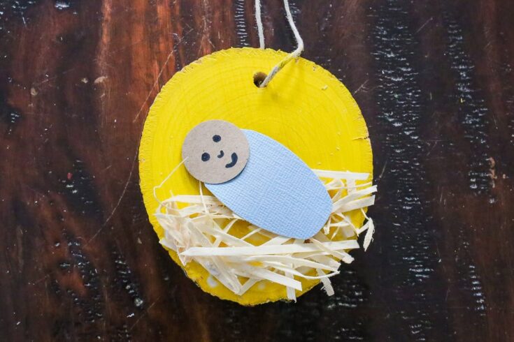 Cute handmade baby Jesus ornament with a smiling face, blue body, and straw bed, hanging on a yellow wooden background, perfect for Christmas decorations.