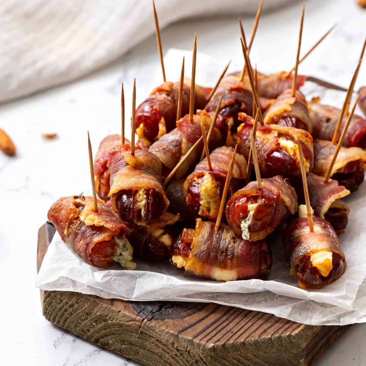 Bacon-wrapped dates stuffed with cheese, secured with toothpicks, served on a rustic wooden board for appetizer or party snack.