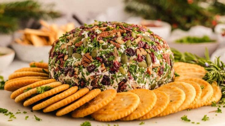 A cheese ball coated with herbs, nuts, and dried fruits, surrounded by round crackers on a festive table setting, perfect for holiday gatherings and celebrations.