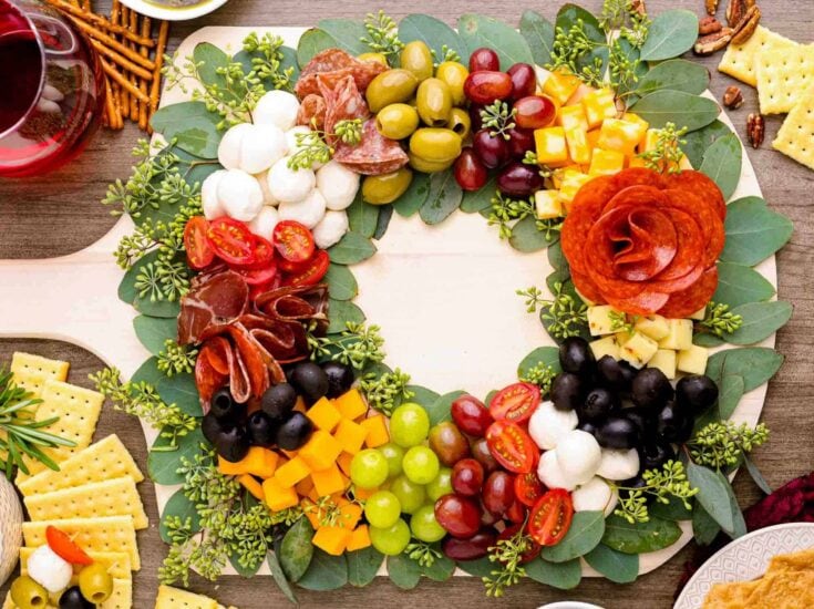 Colorful cheese, grapes, cherry tomatoes, and cured meats arranged on a round platter with greenery for a festive appetizer display.