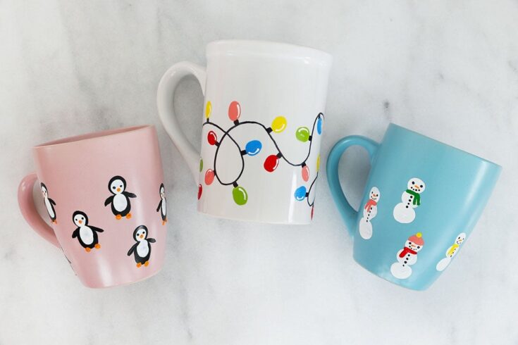 Set of three colorful holiday mugs featuring penguins on a pink mug, snowmen on a blue mug, and Christmas lights on a white mug, perfect for winter celebrations.