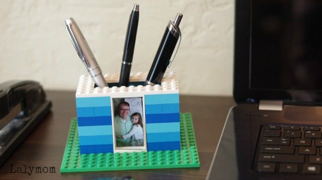 Lego pen holder in blue and green with three pens and a family photo, placed on a desk next to a laptop for a professional workspace setup.