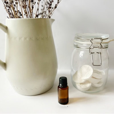 A beige ceramic vase with dried lavender, a small amber essential oil bottle, and a glass jar filled with cotton pads on a white surface, ideal for natural wellness.