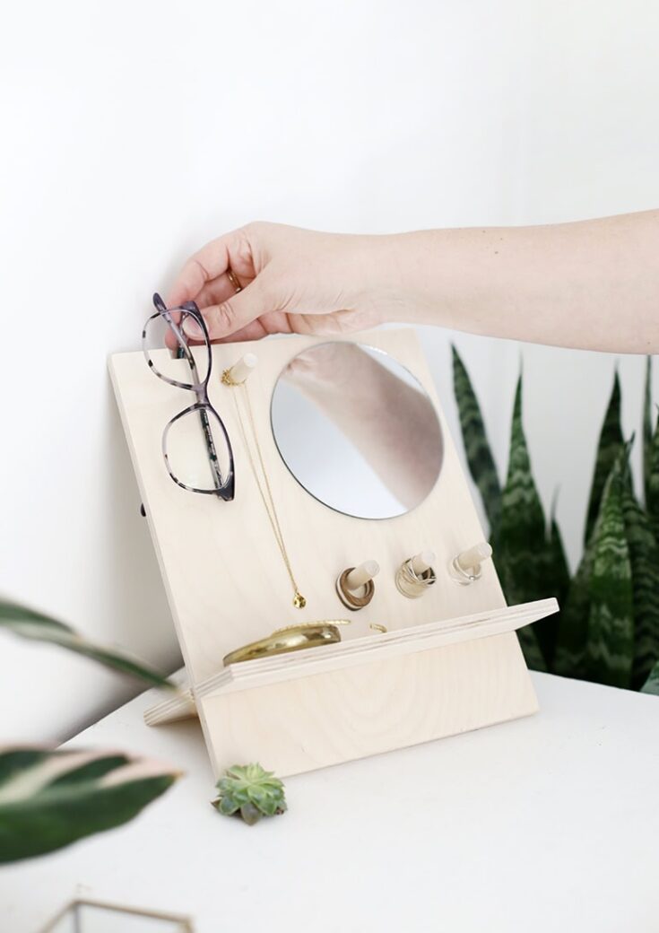 Hand placing glasses on a wooden jewelry organizer with mirror, hooks, and compartments, on a white surface with green plants in the background.