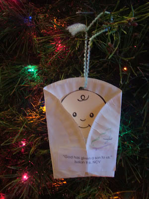 Festive Christmas tree ornament made from a white cloth with a hand-drawn angel face, hanging on a decorated Christmas tree with colorful lights.