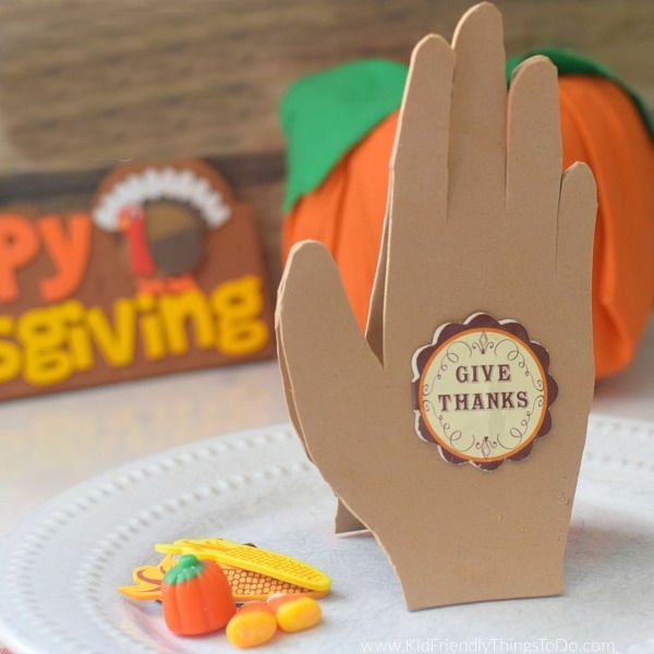 A paper hand card with a 'Give Thanks' sticker, surrounded by miniature pumpkins and fall-themed decorations, celebrating Thanksgiving and autumn.