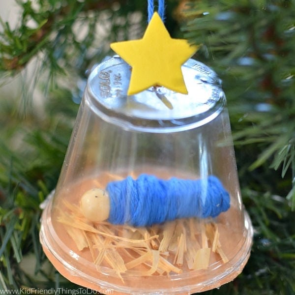 Decorative glass jar with a yellow star on top, containing a blue yarn ball and a small white bead, set among green pine branches for holiday decor.