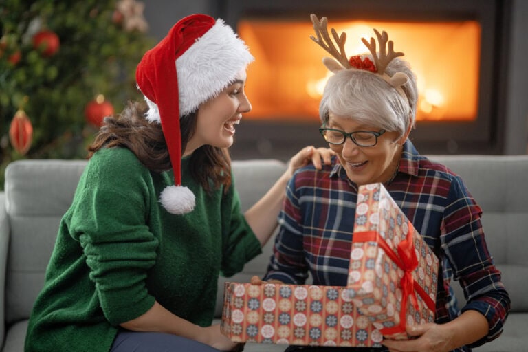 A young woman and an elderly woman exchanging gifts during Christmas, with festive decorations and a cozy fireplace in the background.