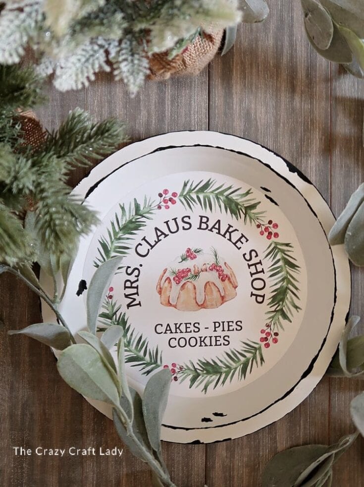 Decorative plate from Mrs. Claus Bake Shop featuring a gingerbread cookie with holly and pine branches, surrounded by festive holiday design, on a rustic wooden table.