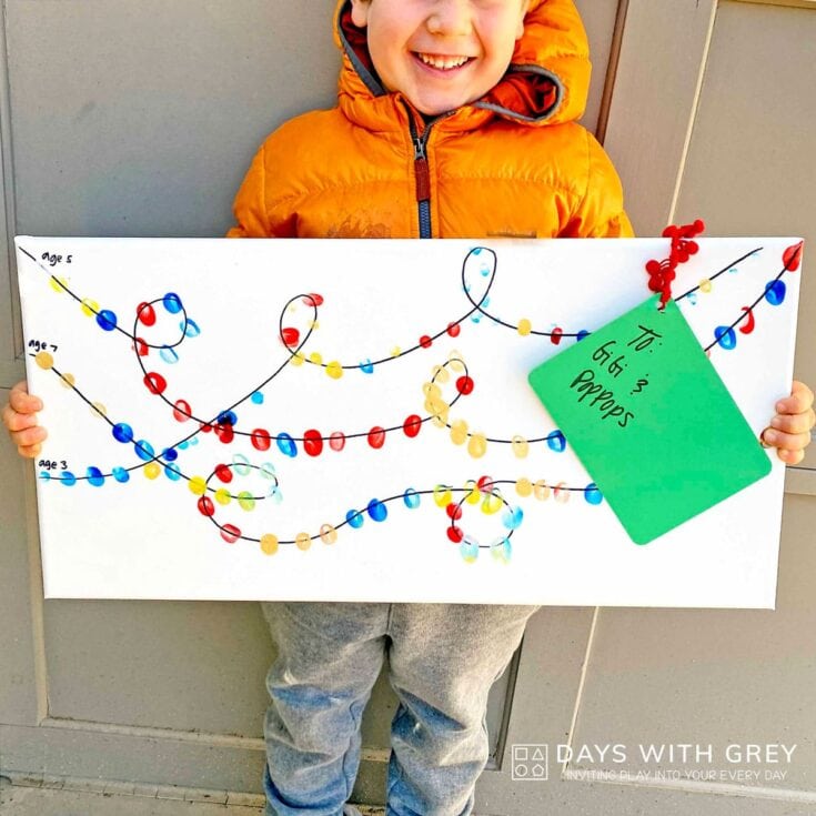 Young child in orange jacket holding a white board with colorful string art designs and a green note attached, celebrating a craft project at age 3.