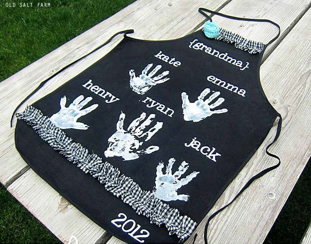 Custom black apron with white handprints and names of family members Kate, Ryan, Henry, Jack, Emma, and Grandma, with the year 2012, on a wooden table outdoors.