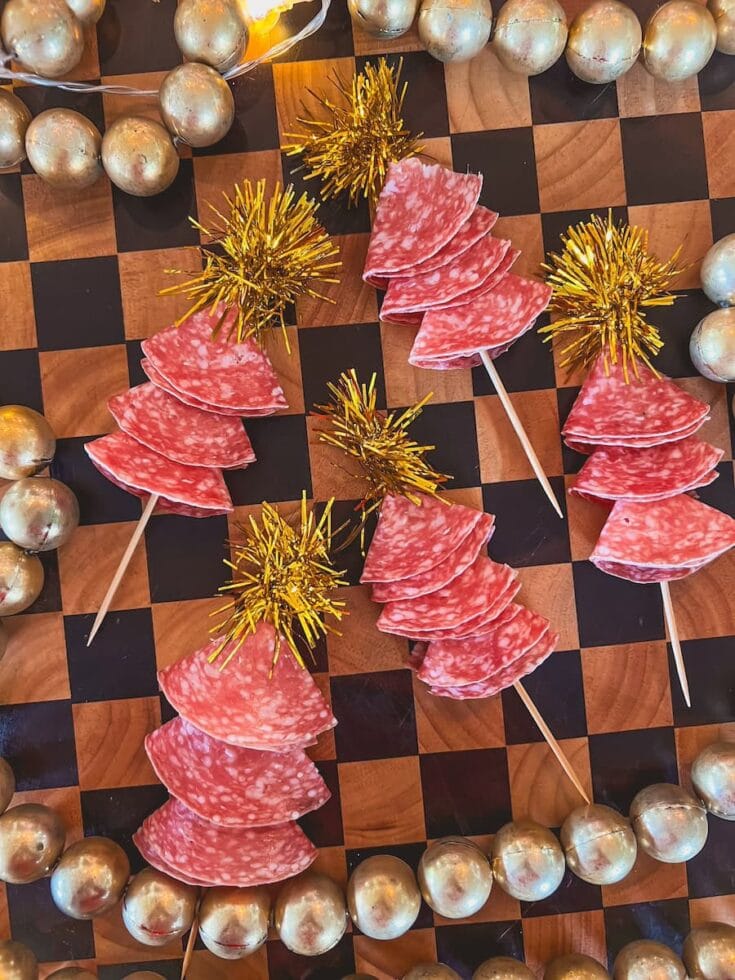 Holiday-themed skewers with slices of salami and gold tinsel on a chessboard background, surrounded by silver ornaments, perfect for Christmas or New Year celebrations.