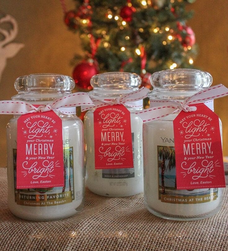 Three white candles in glass jars with red ribbons and Christmas tags, placed in front of a decorated Christmas tree with red ornaments and warm lights.