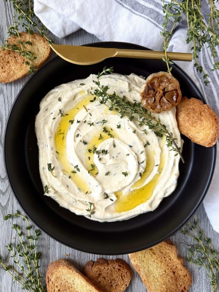 A bowl of creamy hummus garnished with olive topping, fresh thyme, and drizzled with olive oil, served with toasted bread slices and crispy croquettes on a rustic wooden table.