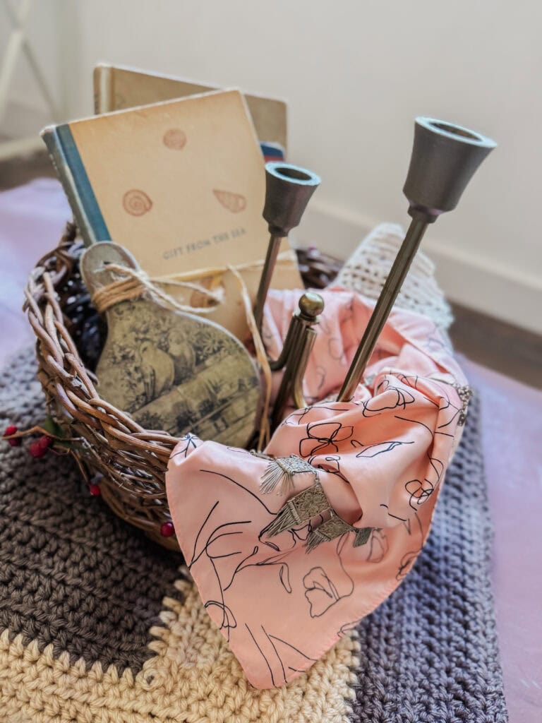 A decorative basket filled with candles, a book, and a plush toy, styled with a pink cloth and placed on a textured surface for home decor.