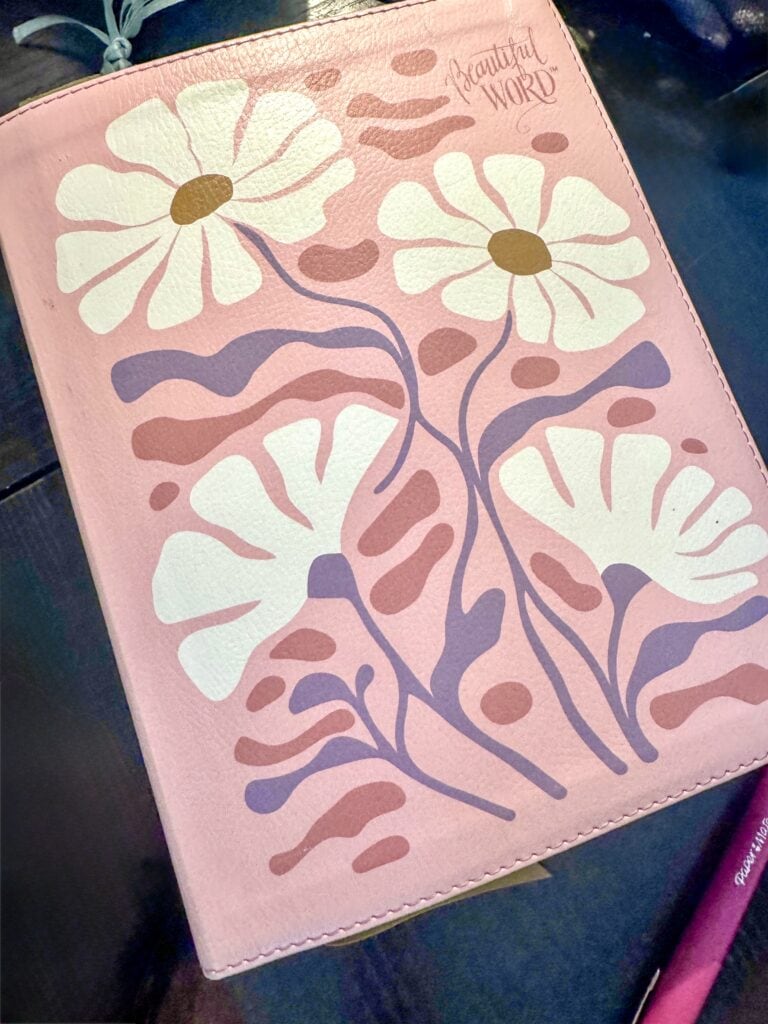 Pink floral notebook with white daisy-like flowers and purple stems, featuring the words 'Beautiful Word' in the top right corner, ideal for stationery and gift ideas.