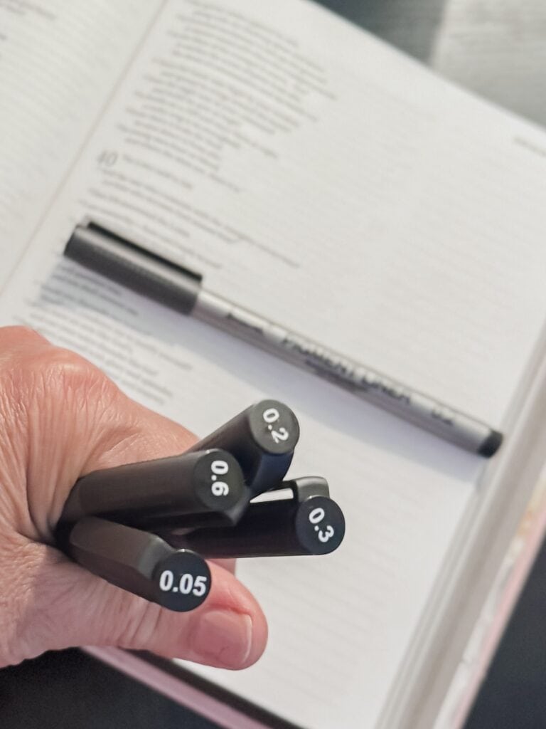 Close-up of a hand holding black markers with different tip sizes over an open notebook with printed text, in a well-lit environment.