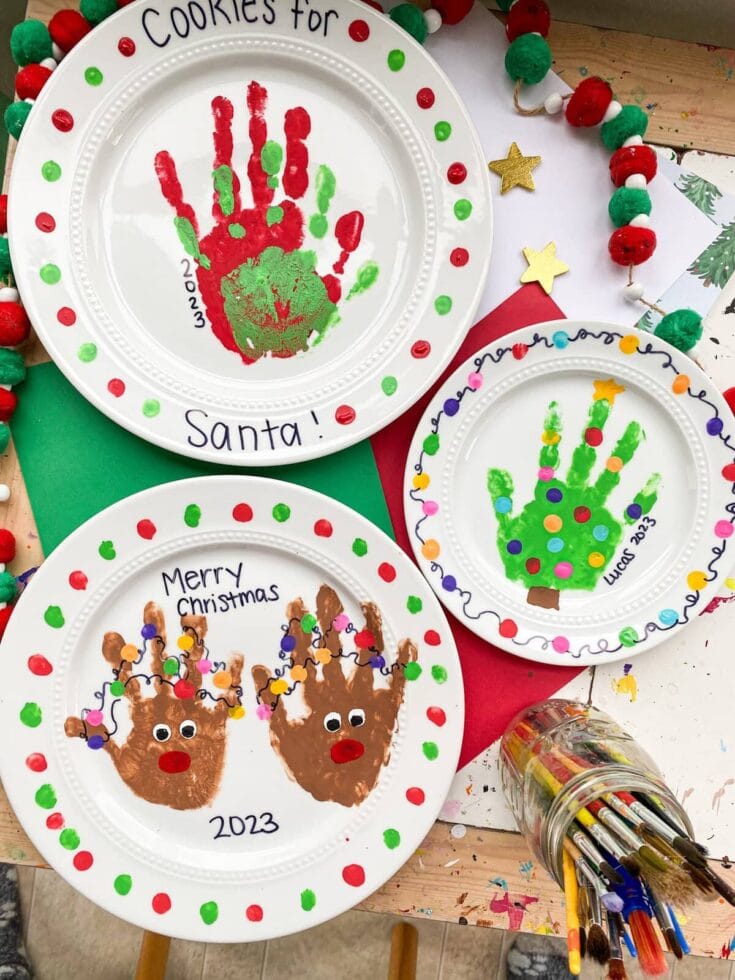 Festive holiday cookie plates featuring handprint art of Santa, reindeer, and Christmas tree, decorated with colorful dots and holiday messages for Christmas 2023.