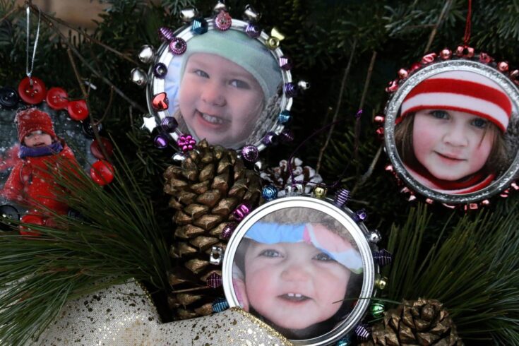 Three children with winter hats and scarves smiling behind decorated mirrors on a Christmas tree, surrounded by pinecones and ornaments, celebrating the holiday season.
