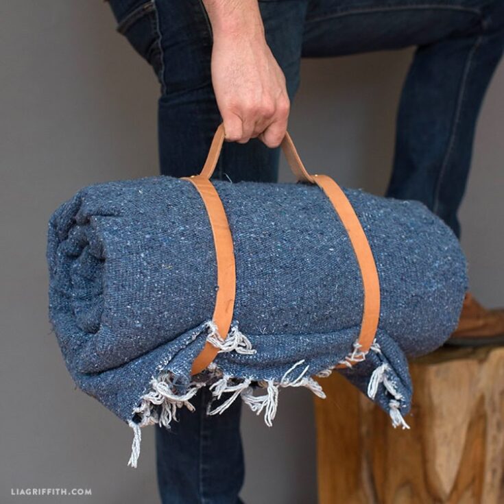 A person holding a rolled blue textured blanket with leather straps, standing next to a wooden stool, showcasing cozy home decor and comfort.