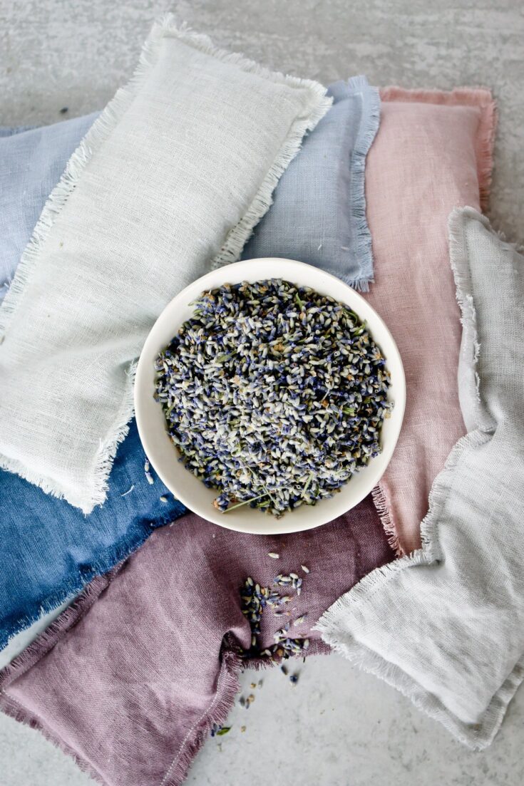 A bowl of dried lavender flowers surrounded by pastel-colored linen fabrics in shades of white, blue, pink, and purple, arranged on a light gray surface.