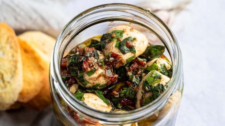 Close-up of herbed mozzarella balls marinated with herbs and sun-dried tomatoes in a glass jar, perfect for antipasto or salads.