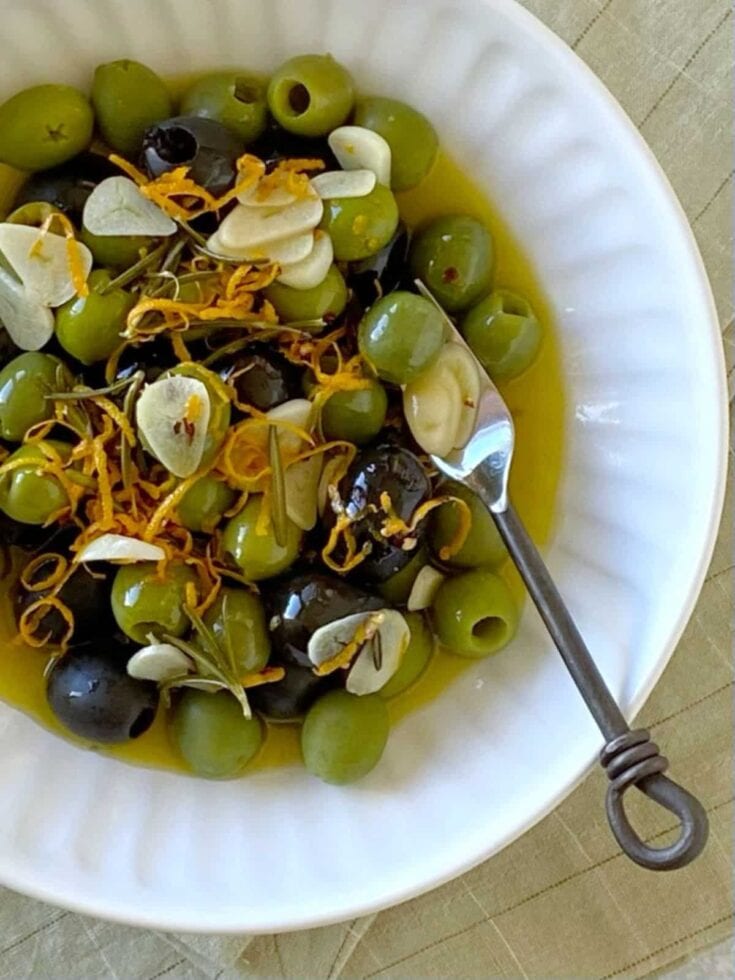 A bowl of green and black olives garnished with orange zest, garlic slices, and herbs, served with a small spoon for tasting, on a white dish.