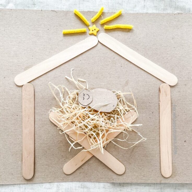 Handmade baby bird nest art using popsicle sticks, shredded paper, and a small wooden bird with a smiling face, arranged to resemble a house with a sun at the top.