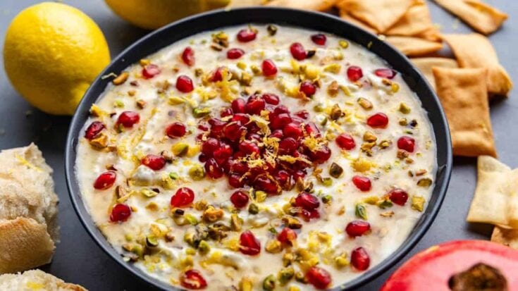Creamy rice pudding garnished with pomegranate seeds and pistachios, served in a black bowl with lemon and pita bread on a dark surface, highlighting Middle Eastern dessert flavors.