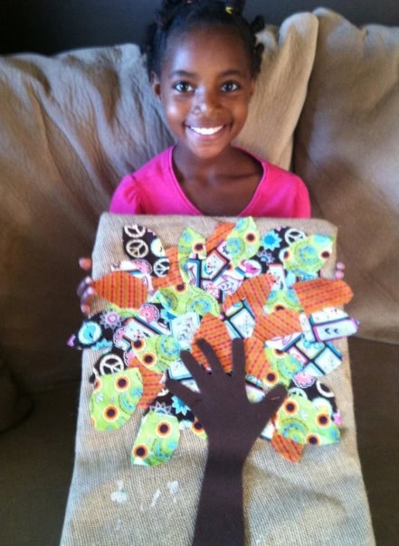 Smiling young girl holding a craft project featuring a black paper tree with colorful butterfly cutouts on a beige background, celebrating creativity and childhood art.