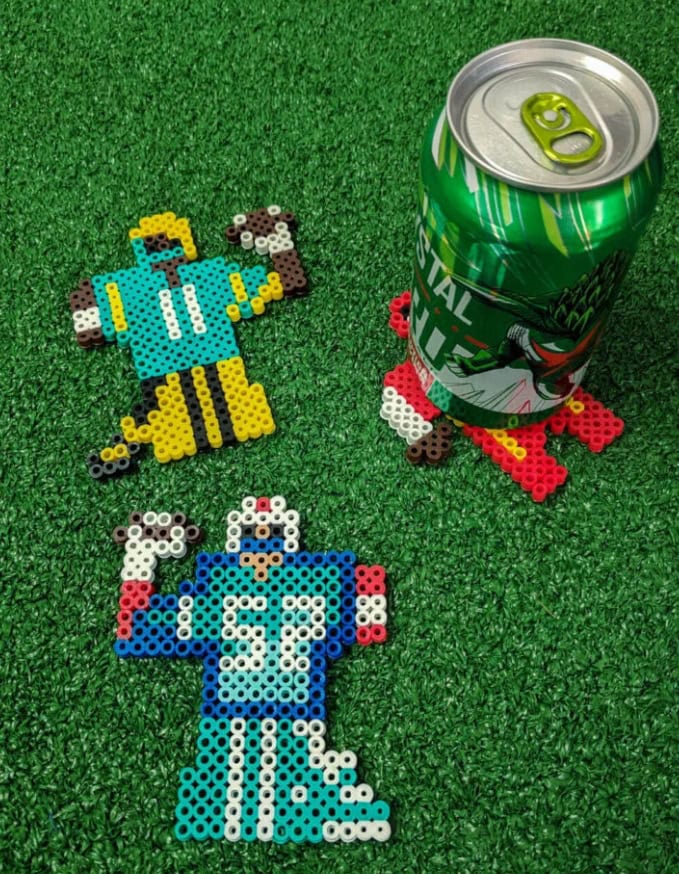 Colorful pixel art bead crafts of two characters on green artificial grass, with a green soda can featuring a cartoon design nearby, showcasing DIY craft ideas.