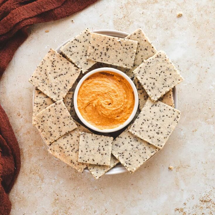 Seed crackers arranged in a circle around a bowl of orange dipping sauce on a light surface, healthy snack presentation.