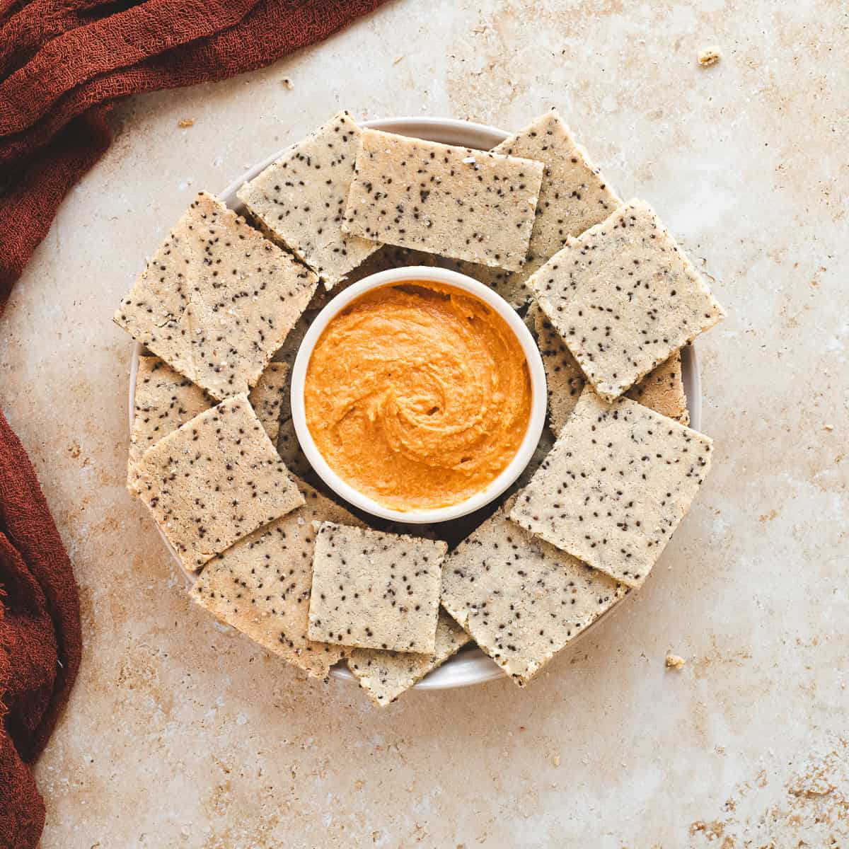 Seed crackers arranged in a circle around a bowl of orange dipping sauce on a light surface, healthy snack presentation.