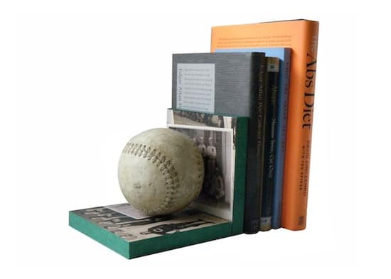 A baseball resting on a shelf with a collection of books behind it, including titles on art, design, and literature, arranged in a modern, minimalist style.