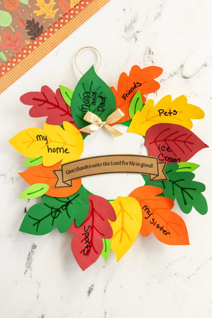 Colorful paper leaf wreath with handwritten messages like 'my home,' 'pets,' 'friends,' and 'my sister,' decorated with a ribbon and a thankfulness message, on a white background.