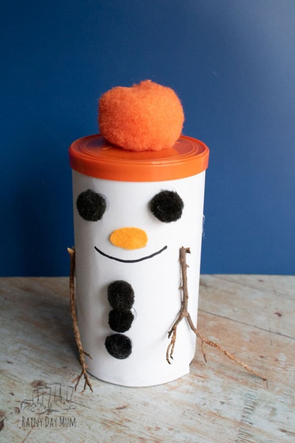 Creative snowman craft made from a white container with black buttons, orange nose, twig arms, and a fuzzy orange pom-pom hat, perfect for holiday decorations.