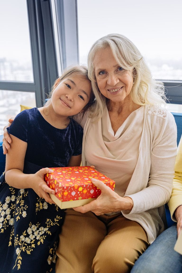 Grandmother and young girl sharing a joyful moment while exchanging a wrapped gift in a bright, modern room with large windows and city view.