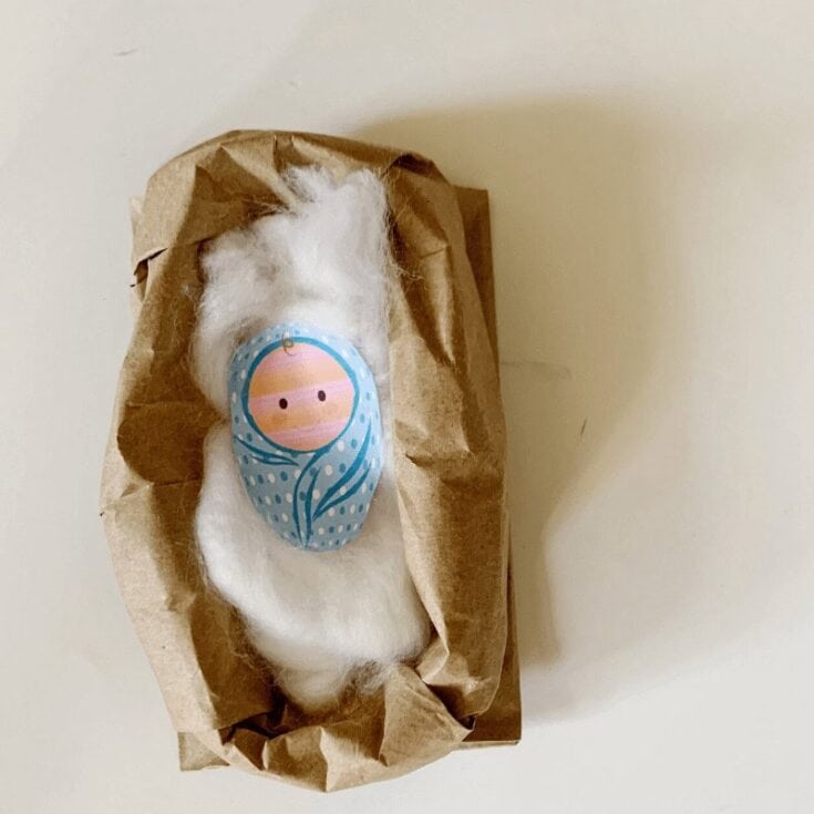 A handmade wool doll with a blue and pink face, wrapped in brown paper, on a plain white background, showcasing artisanal craft and textile art.