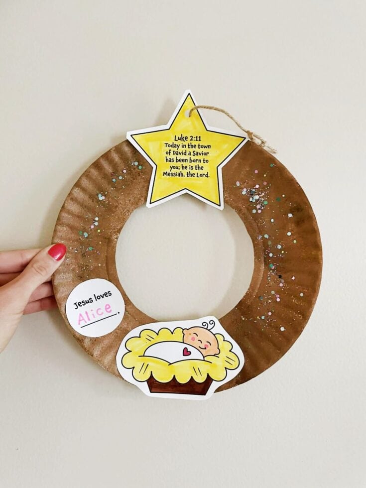 Hand holding a brown paper advent wreath decorated with stickers including a star with Bible verse Luke 2:11, a sleeping baby Jesus, and a message Jesus loves Alice, for Christmas celebration.