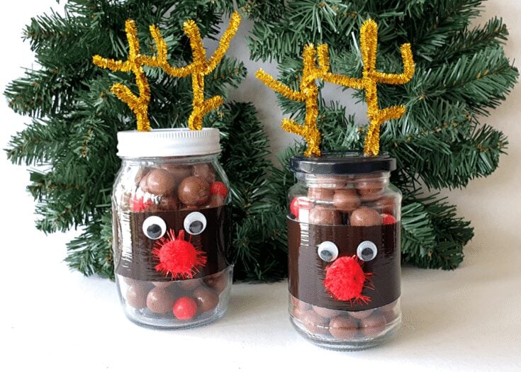 Two mason jars decorated as reindeer with googly eyes, red pom-pom noses, and gold pipe cleaner antlers, set against a green Christmas tree background for holiday decor.